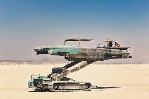 A unique, art-car vehicle with treads sits on the dusty playa under a clear blue sky.