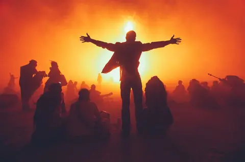 A silhouetted figure with arms outstretched stands before a blazing sunset at a festival.