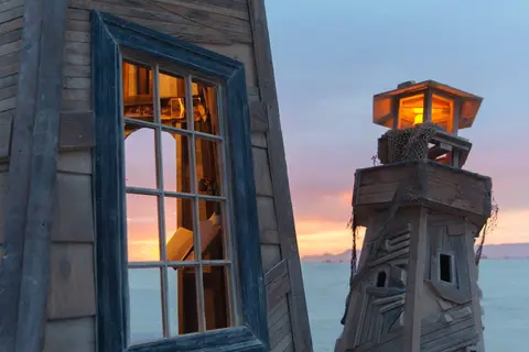 A rustic, weathered lighthouse structure with a glowing lantern room stands against a soft pastel sunset over the water.