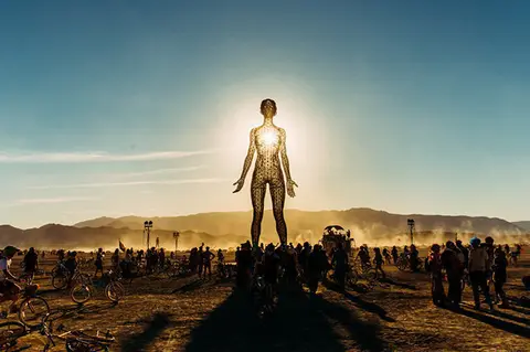 A silhouetted figure stands illuminated by the sun at the center of a vast desert gathering.