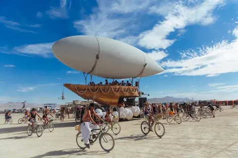 A massive blimp-like art installation floats over a dusty desert landscape filled with people on bicycles.