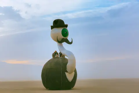 A whimsical, cartoonish bird sculpture with a bowler hat and mustache sits under a vast, cloudy sky.