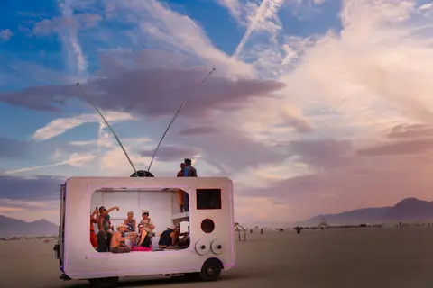 A vintage television art installation with people inside sits in the vast desert landscape under a dramatic sky.