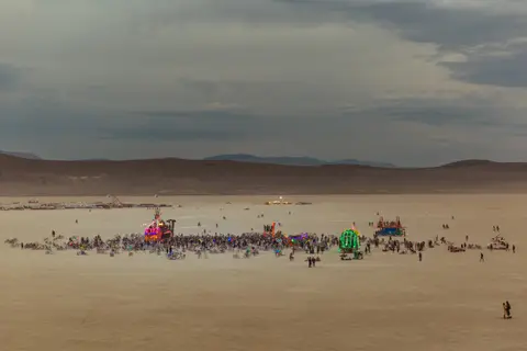 A wide shot of a festival crowd gathered around art installations on a vast, dusty plain under a cloudy sky.