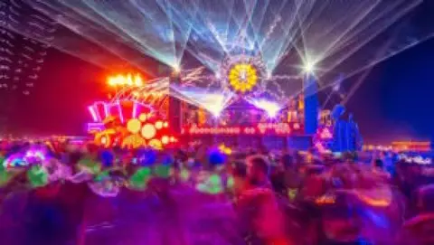 A vibrant, laser-lit stage pulses with energy above a blurred crowd at a nighttime music festival.