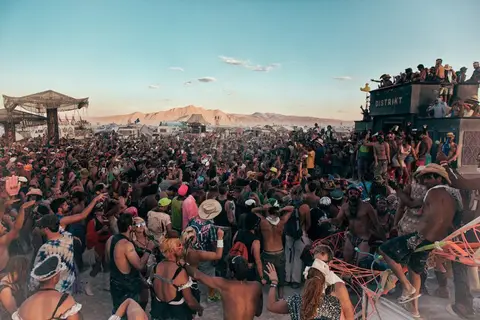 A massive crowd gathers at a desert festival under a vast, clear sky with mountains in the distance.