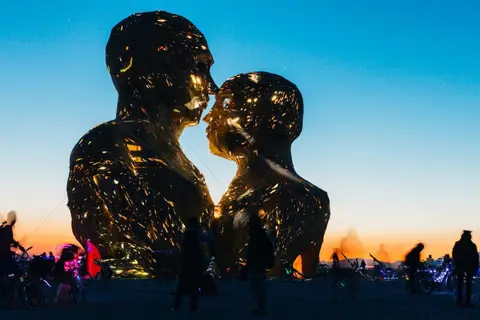 Two illuminated figures embrace against a vibrant desert sunset sky at a festival.