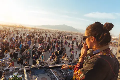A DJ plays to a massive crowd at a desert festival during golden hour.