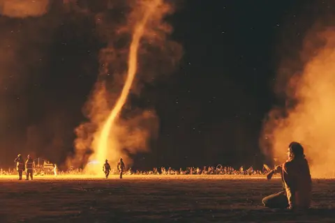 A lone figure sits in the foreground watching a massive fire tornado erupt against the night sky at a festival.