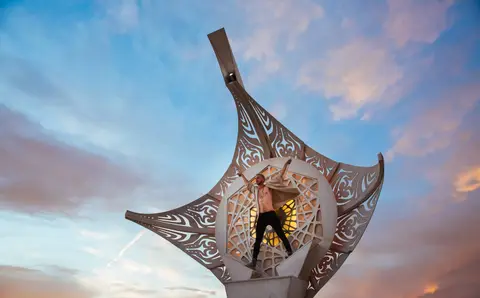 A person stands with arms outstretched inside a large, ornate, metallic sculpture against a dramatic sunset sky.