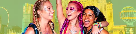 Three young women with glitter and colorful hair smile and laugh together at a vibrant outdoor event.