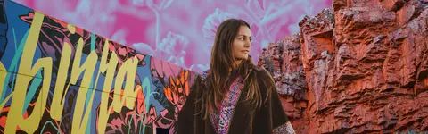 A woman gazes off to the side, framed by vibrant graffiti art on the left and rugged, sun-drenched rock formations on the right.