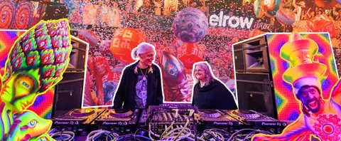 Two DJs smile behind their turntables at a vibrant, psychedelic-themed festival stage with colorful decorations and a crowd in the backgroun