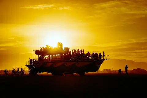 Silhouetted figures stand atop a large, decorated vehicle against a vibrant, hazy sunset at a festival.
