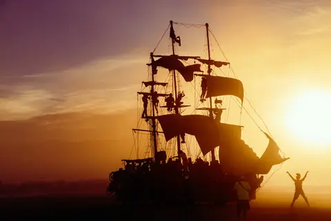 Silhouette of a pirate ship with people on deck against a vibrant sunset sky, with a lone figure celebrating on the right.