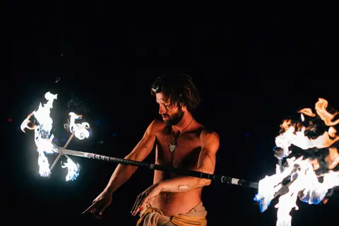 A shirtless man spins a fiery staff against a dark background.