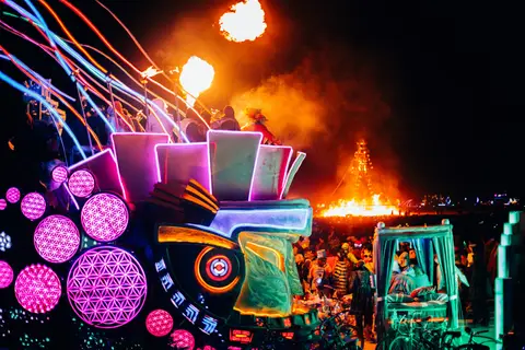 A vibrant night scene at Burning Man features illuminated art installations and a large bonfire in the background.