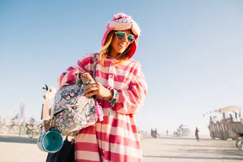 A woman in a pink and white checkered onesie and fuzzy hat smiles while holding a backpack at Burning Man.