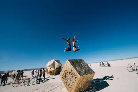 Two people joyfully leap into the air against a clear blue sky at a desert festival, with unique art installations in the foreground and bac