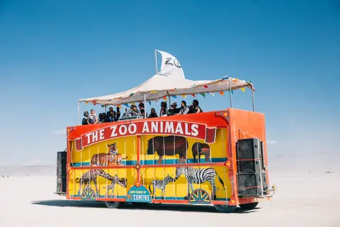 A brightly painted bus, labeled 'The Zoo Animals', carries passengers across a vast, flat, sun-drenched playa under a clear blue sky.
