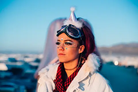 A woman with festival makeup and goggles looks off into the distance under a clear blue sky.