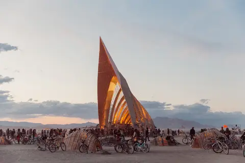 A striking, angular art installation stands tall on the playa at sunset, surrounded by a crowd and bicycles.