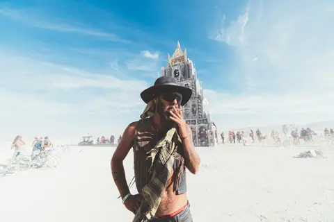 A man in a hat and vest smokes a cigarette in front of the Man structure at Burning Man.