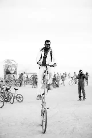 A man rides a tall bicycle through a dusty, outdoor gathering with many people and bicycles in the background.