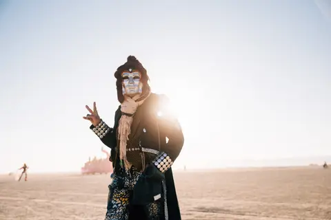 A person in elaborate costume and face paint flashes a peace sign against a bright, sunny desert sky.