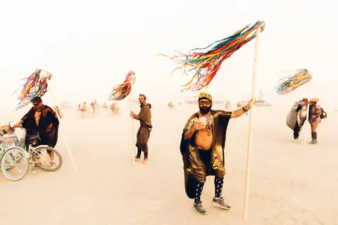 A man with a colorful streamer he is holding aloft stands in a dusty, open field with other people and art installations in the background.