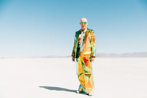 A person in a vibrant, colorful suit stands in a vast, flat, white desert under a clear blue sky.