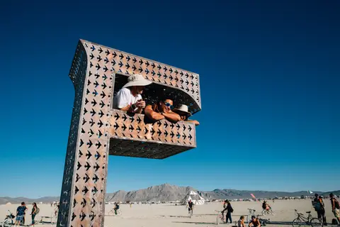 People observe the desert landscape from within a large, art installation structure under a clear blue sky.