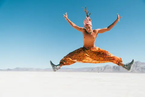 A man in a turban and patterned pants leaps in the air with arms outstretched against a clear blue sky.