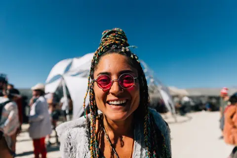 A smiling woman with braided hair and pink sunglasses poses for a photo at an outdoor event.