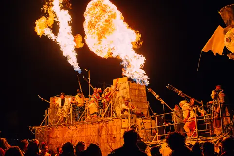 A large, fiery art installation erupts with flames against the night sky at a festival.