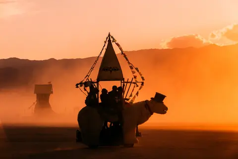 A whimsical art car shaped like a camel carries silhouetted figures across a dusty desert landscape at sunset.