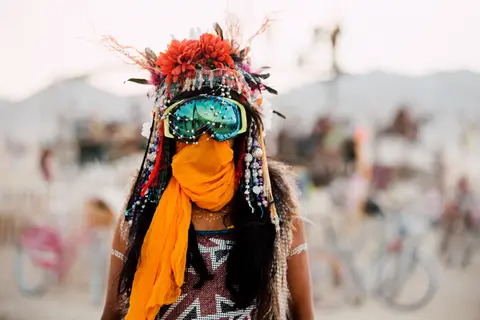 A person in elaborate headdress and goggles stands at a festival, their face obscured by a bright orange scarf.