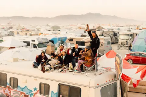 A diverse group of people pose joyfully on top of an RV at a desert festival.
