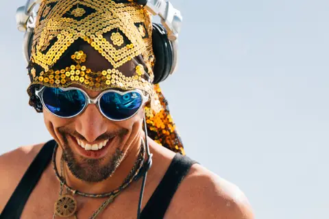 A smiling man with a gold sequined bandana and heart-shaped sunglasses wears headphones.