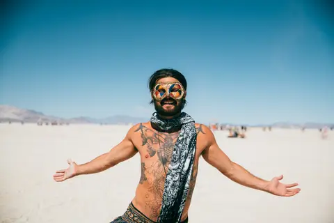 A man with a beard, tattoos, and colorful goggles around his neck, stands with arms outstretched in a vast, flat, dusty landscape under a cl