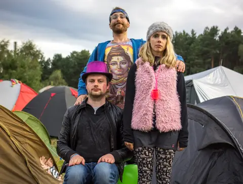 Three friends pose for a photo in a tent village at an outdoor festival.