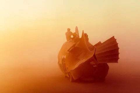 Silhouetted figures stand atop a whimsical, futuristic sculpture against a hazy, golden sunset.