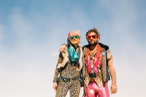 A vibrant couple poses against a clear blue sky, dressed in flamboyant festival attire.