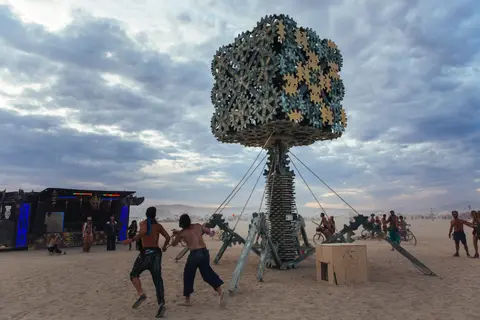 Two figures dance in the foreground of a large, metallic tree sculpture at a desert festival.