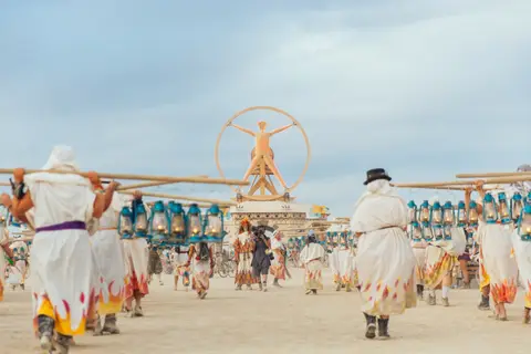 A procession of people in white robes carries a structure towards the iconic Man statue at Burning Man.