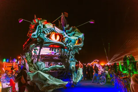 A fantastical, illuminated dragon-like art car roars to life at night amidst a crowd of festival-goers.