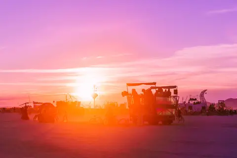 A vibrant sunset paints the sky in hues of purple and orange over a dusty desert landscape with a large, decorated vehicle carrying people.