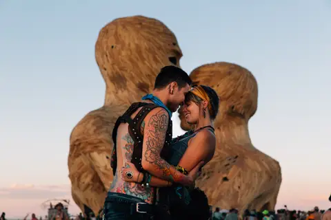 A couple embraces tenderly in front of a large, abstract sculpture at sunset.