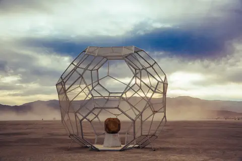 A geodesic dome art installation sits in the vast, dusty desert landscape under a dramatic cloudy sky.