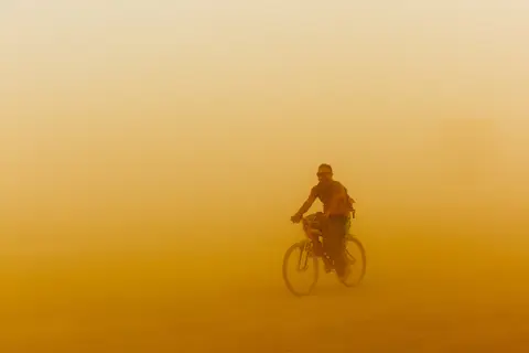 A lone cyclist rides through a dense, orange dust storm.